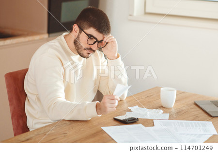 A man is seated at table, concentrating on papers in his hand with a thoughtful expression. Scattered documents, a calculator on the table, indicating a session of financial review or budgeting A man is seated at table, concentrating on papers in his hand with a thoughtful expression. Scattered documents, a calculator on the table, indicating a session of financial review or budgeting 114701249