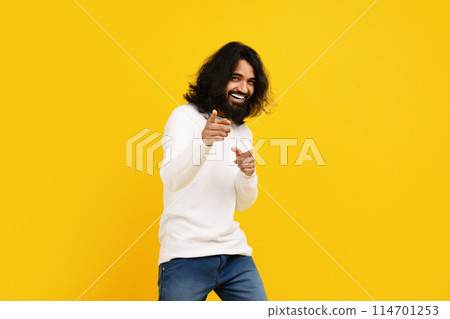 Indian man with long hair and a beard is standing in front of a bright yellow background, pointing at camera and smiling Indian man with long hair and a beard is standing in front of a bright yellow background, pointing at camera and smiling 114701253