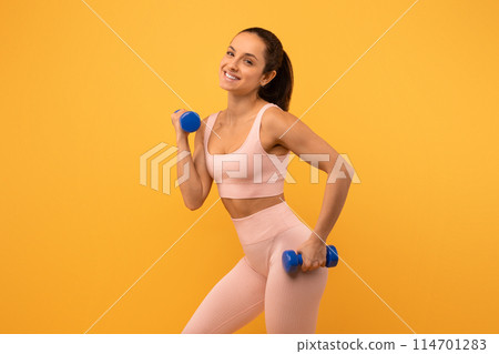 A woman with long hair wearing a pink sports bra is standing with both hands holding two blue dumbbells. She appears to be in a gym environment, possibly working out. 114701283