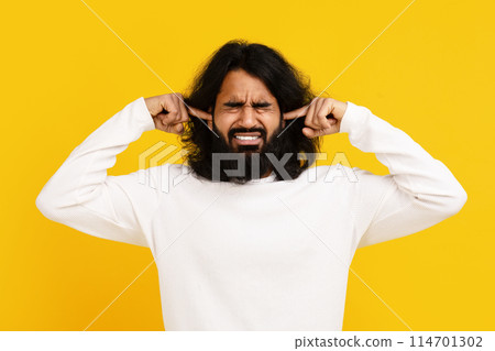 Indian man with long hair and a beard is shown holding his hands on his ears, indicating a gesture of blocking out noise or protecting his ears from loud sounds 114701302