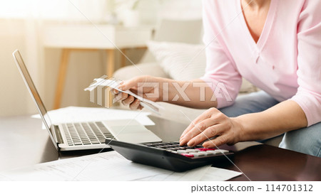 Cropped of woman is seated at a desk, focused on her work. She is using a calculator and a laptop to perform financial calculations or analysis. The setting is a typical office environment. 114701312