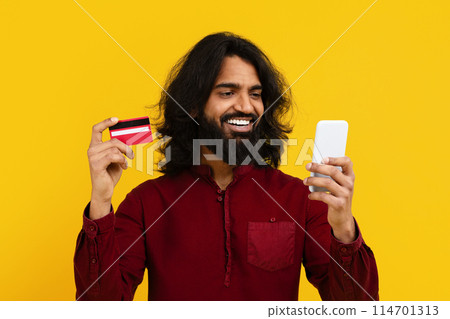 Indian man is shown holding a smart phone in one hand and a credit card in the other. He appears to be making a transaction or online purchase using the two devices. 114701313