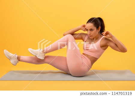 A focused young woman in a pink athletic outfit is engaged in a fitness routine, executing bicycle crunches on a blue yoga mat, vivid yellow backdrop that highlights her exercise activity. 114701319