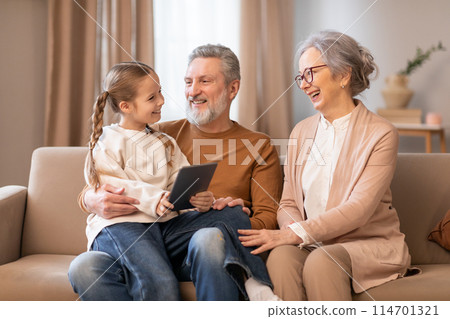An elderly couple and their young granddaughter are seated together on a beige couch, sharing a moment of joy and connection. The grandfather holds a digital tablet 114701321