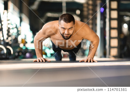 Shirtless man is performing push ups in a gym, his muscles strained as he lowers and raises his body. The gym equipment and other people working out in the background add to the active atmosphere. Shirtless man is performing push ups in a gym, his muscles strained as he lowers and raises his body. The gym equipment and other people working out in the background add to the active atmosphere. 114701381