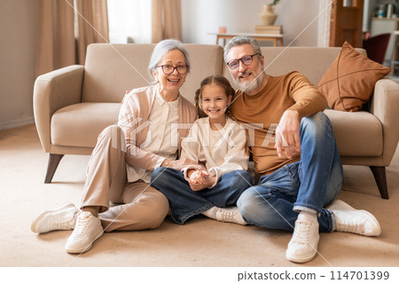 A cheerful elderly couple and their young granddaughter are sitting closely together on the floor of a cozy living room, projecting warmth and family togetherness. A cheerful elderly couple and their young granddaughter are sitting closely together on the floor of a cozy living room, projecting warmth and family togetherness. 114701399