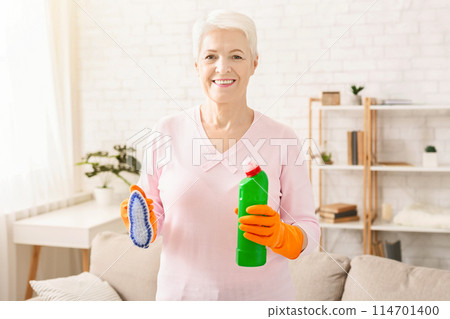 Senior woman is shown holding a bottle of cleaner and a rag in her hands. She appears to be ready to clean or wipe surfaces, displaying household chores in action. Senior woman is shown holding a bottle of cleaner and a rag in her hands. She appears to be ready to clean or wipe surfaces, displaying household chores in action. 114701400