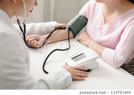 A doctor is using a stethoscope to measure a patients blood pressure in a medical setting. The doctor is focused on listening to the patients heartbeat and recording the readings on a medical chart. A doctor is using a stethoscope to measure a patients blood pressure in a medical setting. The doctor is focused on listening to the patients heartbeat and recording the readings on a medical chart. 114701404