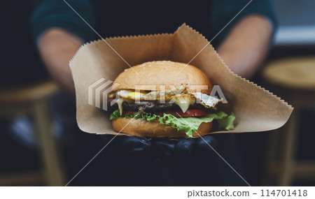 Fresh burger cooked at barbecue outdoors in craft paper. Cookout american bbq food. Big hamburger with steak meat and vegetables closeup with chef unfocused at background. Street food, fast food. 114701418