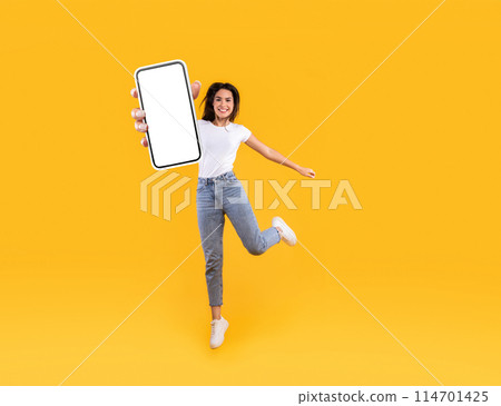 Happy young woman showing empty cellphone screen while jumping up on yellow studio background, collage, full body length, banner panorama. Excited lady recommending nice and useful mobile application 114701425