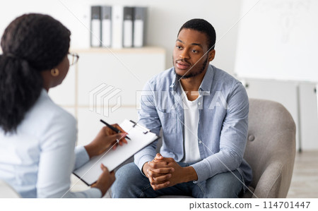 Unhappy young black man having session with professional psychologist at mental health clinic. Psychotherapist taking notes during conversation with depressed male patient Unhappy young black man having session with professional psychologist at mental health clinic. Psychotherapist taking notes during conversation with depressed male patient 114701447