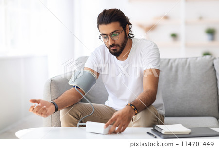 Young arab man sitting on couch, checking blood pressure and taking notes at home. Indian guy in white t-shirt using modern tonometer, experiencing hypertension caused by stress, copy space Young arab man sitting on couch, checking blood pressure and taking notes at home. Indian guy in white t-shirt using modern tonometer, experiencing hypertension caused by stress, copy space 114701449