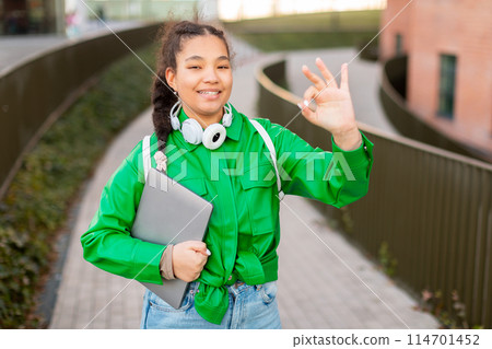 A cheerful young girl with her hair in braids is standing on a city sidewalk, flashing okay gesture with her right hand, holding a laptop, on her way to an activity or school. A cheerful young girl with her hair in braids is standing on a city sidewalk, flashing okay gesture with her right hand, holding a laptop, on her way to an activity or school. 114701452