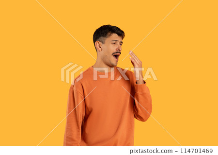 A young man with dark hair stands against a vivid yellow backdrop, covering his mouth with his hand as he yawns widely, displaying signs of fatigue or boredom wearing a casual orange sweatshirt. 114701469