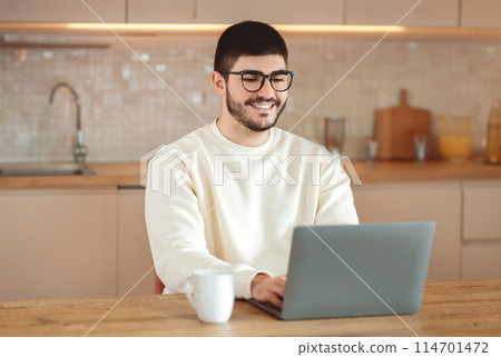 A man is sitting at a table, focused on using his laptop computer. He appears to be working or studying intently, typing and clicking on the laptop screen. The background is simple and nondescript. 114701472