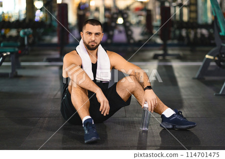 A man in athletic clothing sitting on the floor of a gym, surrounded by exercise equipment. He appears to be taking a break or stretching after a workout. 114701475