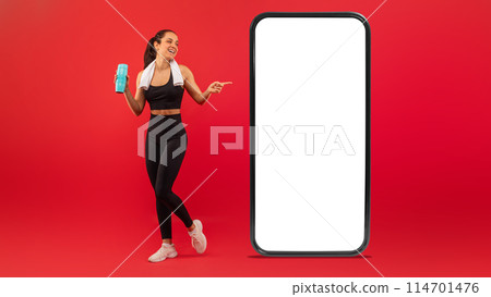 A smiling young woman dressed in black sportswear stands confidently next to an oversized representation of a smartphone with a blank screen, holding a bottle of water 114701476