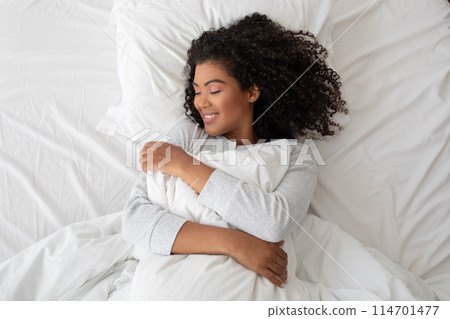Hispanic woman with curly hair smiles contentedly as she embraces a fluffy pillow, lying in comfort on a bright white bedspread Hispanic woman with curly hair smiles contentedly as she embraces a fluffy pillow, lying in comfort on a bright white bedspread 114701477
