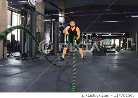 A man is working out in a gym, holding a rope and engaging in various exercises to strengthen his upper body and core muscles, focused and determined on the intense workout routine. 114701503