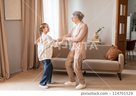 An older woman and a young girl are dancing in a cozy living room. The woman is wearing glasses and a floral dress while the girl is in casual clothes 114701529