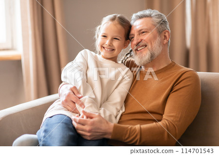 Elderly man and a little girl are seated on a couch. Grandfather appears to be talking to the girl while she listens attentively. They seem to be engaged in a conversation or storytelling. 114701543