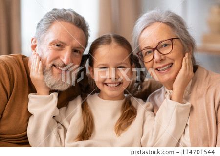 A warm and affectionate moment as a grey-haired grandfather and grandmother sit closely with their beaming young granddaughter, looking directly at the camera 114701544