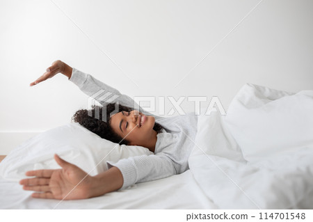 Hispanic woman is lying in bed with her arms stretched out to the sides. She appears relaxed and comfortable, possibly waking up or winding down for the day. Hispanic woman is lying in bed with her arms stretched out to the sides. She appears relaxed and comfortable, possibly waking up or winding down for the day. 114701548