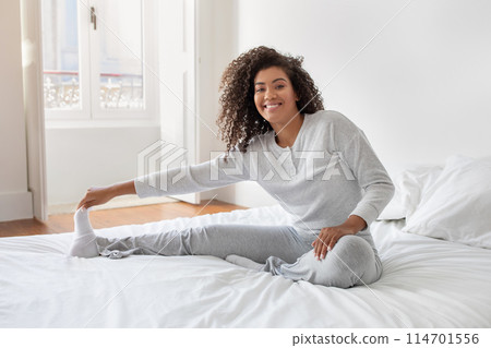 A cheerful Hispanic woman with curly hair is sitting on a neatly made bed in a bright bedroom. She is wearing a cozy white sweater and gray pants A cheerful Hispanic woman with curly hair is sitting on a neatly made bed in a bright bedroom. She is wearing a cozy white sweater and gray pants 114701556