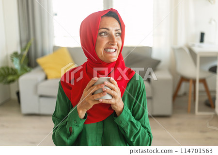 A cheerful woman wearing a red hijab and green blouse stands in a sunlit living room, holding a gray mug with both hands. She gazes out of the frame with a joyful expression 114701563