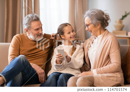 A family of three individuals, consisting of grandparents and little girl, sitting together on a couch in a living room. They are focused on a cell phone screen A family of three individuals, consisting of grandparents and little girl, sitting together on a couch in a living room. They are focused on a cell phone screen 114701574