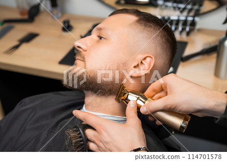Barber skillfully uses a trimmer to shape the clients beard, focusing on the contours of the cheek. Barber skillfully uses a trimmer to shape the clients beard, focusing on the contours of the cheek. 114701578