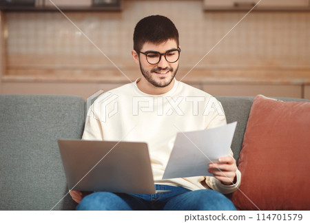 A man is seated on a couch with notebook on his lap, focused on a piece of paper in his hands, entrepreneur working from home A man is seated on a couch with notebook on his lap, focused on a piece of paper in his hands, entrepreneur working from home 114701579