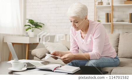 A senior woman with short white hair is seated at a cozy living space, meticulously examining financial papers. She appears concentrated, with a calculator, notebook, and laptop on the table A senior woman with short white hair is seated at a cozy living space, meticulously examining financial papers. She appears concentrated, with a calculator, notebook, and laptop on the table 114701582
