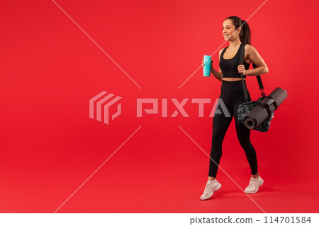 A woman wearing a black sports bra top and leggings is pictured holding a blue cup in her hand, walking towards copy space, isolated on red background A woman wearing a black sports bra top and leggings is pictured holding a blue cup in her hand, walking towards copy space, isolated on red background 114701584