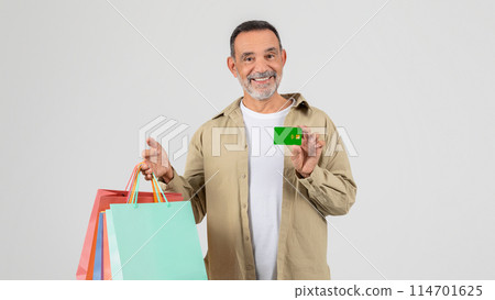 A senior man standing holding shopping bags in one hand and a credit card in the other. He appears to be in a shopping area, possibly a mall, ready to make purchases. 114701625