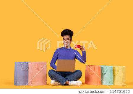 A cheerful young Hispanic woman sits cross-legged between colorful shopping bags, proudly displaying a credit card while looking at an open laptop, suggesting a successful online shopping session A cheerful young Hispanic woman sits cross-legged between colorful shopping bags, proudly displaying a credit card while looking at an open laptop, suggesting a successful online shopping session 114701633