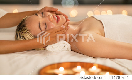 Blonde lady is lying down with her eyes closed, enjoying a gentle facial massage provided by a skilled therapist, side view 114701635