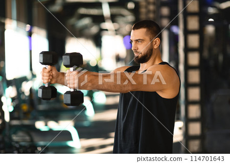 A man is shown holding a pair of dumbbells while standing in a gym. The man appears focused on his workout, showcasing strength and determination in his posture. 114701643