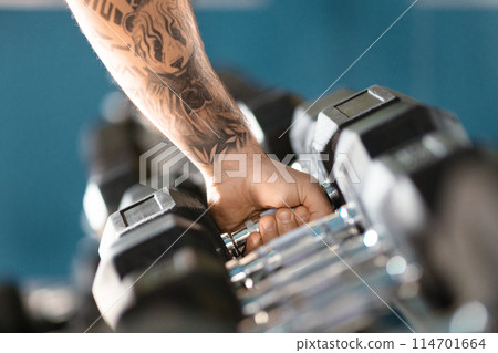 A man with a tattoo on his arm is seen holding a barbell. The mans muscles are visibly tense as he grips the weight. The tattoo on his arm adds a unique element to his appearance, cropped shot 114701664