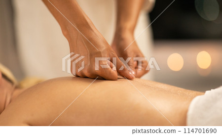 A woman lying face down on a massage table at a spa while a massage therapist performs a back massage. The therapists hands are visible as they apply pressure, closeup 114701690