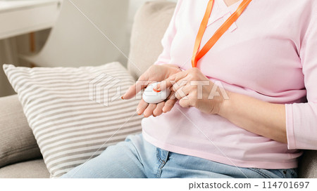 Cropped of elderly woman discreetly holding a white panic button attached to an orange lanyard, suggesting readiness for emergency assistance while she sits comfortably on a cushioned couch 114701697
