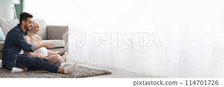 Middle eastern pregnant couple lounges comfortably while using a tablet together, with plenty of copy space for web-banner design on the side 114701726