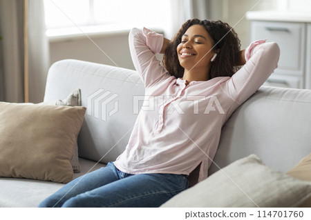 A peaceful scene with a young African American woman sitting comfortably on her couch, wearing earphones and enjoying music, surrounded by a cozy, stylish living room A peaceful scene with a young African American woman sitting comfortably on her couch, wearing earphones and enjoying music, surrounded by a cozy, stylish living room 114701760