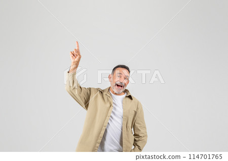 A senior man standing in a tan jacket and white shirt with a neutral expression on his face, pointing finger up at copy space 114701765