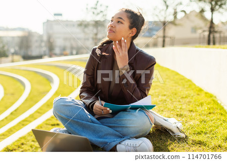 A girl student sitting on the ground outdoors, using a laptop. She appears focused on the screen, typing and browsing. The setting is casual and relaxed, copy space 114701766