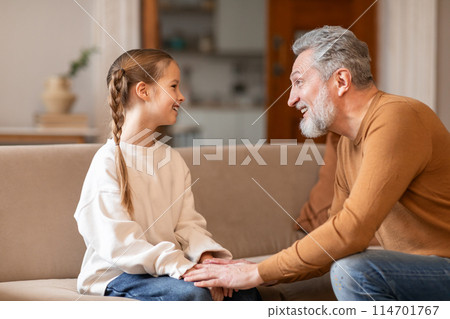 Senior man and a little girl are seated together on a couch in a living room. Grandfather appears to be talking to the girl, who is listening intently 114701767