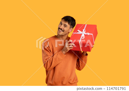A man is holding a red gift box with a white ribbon in his hands. He seems to be preparing to give the gift to someone special, showcasing thoughtfulness and generosity. 114701791