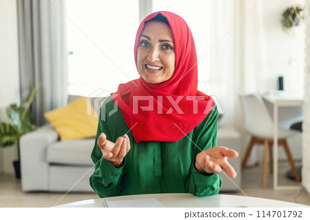 A cheerful woman wearing a bright red hijab and green shirt is engaging in conversation with expressive hand gestures. She is sitting in a well-lit, modern room, possibly her home A cheerful woman wearing a bright red hijab and green shirt is engaging in conversation with expressive hand gestures. She is sitting in a well-lit, modern room, possibly her home 114701792