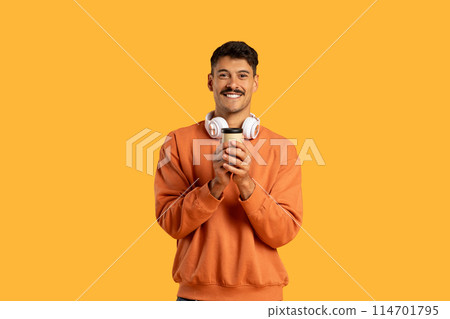 A cheerful man wearing a casual orange sweatshirt stands against a vivid yellow backdrop, holding a white cup in his hands with wireless headphones around his neck 114701795