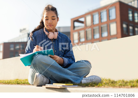 A young girl is seated on the ground, engrossed in a book and wearing headphones. She appears focused and relaxed as she enjoys her reading and music. A young girl is seated on the ground, engrossed in a book and wearing headphones. She appears focused and relaxed as she enjoys her reading and music. 114701806